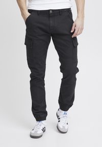Uomo che indossa pantaloni cargo neri con polsini elasticizzati e scarpe da ginnastica bianche con strisce nere, in piedi con una mano in tasca.