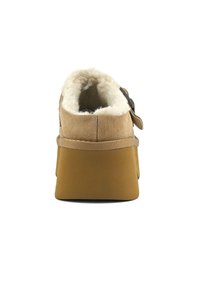 Chausson en suède tan avec doublure blanche douce, semelle en coin basse et sangle latérale ajustable. Vue de dos mettant en valeur la texture douce et la hauteur.