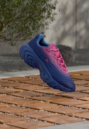 Blå og lyserød sneakers med tekstureret sål svæver over en træ- og stenoverflade udendørs nær et sløret træ og en betonmur.