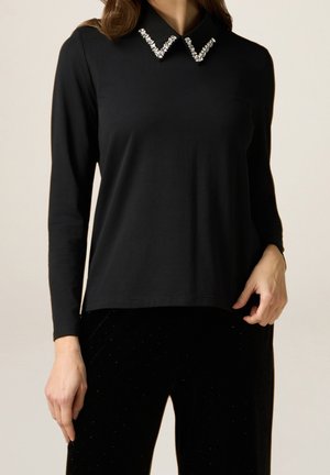 Kobieta ubrana w czarną bluzkę z długim rękawem z ozdobami w kształcie litery V na kołnierzu, zestawioną z czarnymi spodniami o subtelnym kropkowanym wzorze.