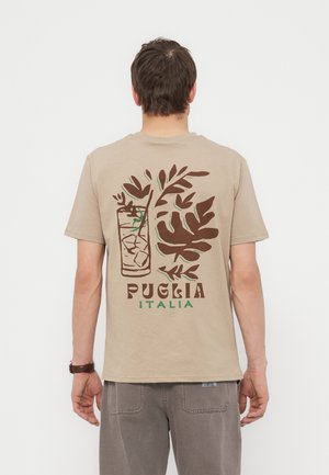 Άνδρας που φοράει μπεζ μπλουζάκι με καφέ και πράσινο γραφικό ποτού με πάγο και φύλλα, κείμενο «Puglia Italia» στην πλάτη.