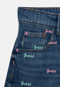 Nærbillede af en lomme på blå denimjeans med orange syninger, pink nitter og flerfarvede broderede "Guess" logoer fordelt over stoffet.