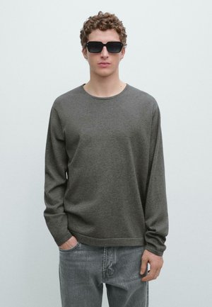 Ung mand med krøllet hår iført sorte solbriller, en mørkegrå langærmet skjorte og falmede grå jeans mod en ensfarvet lys baggrund.