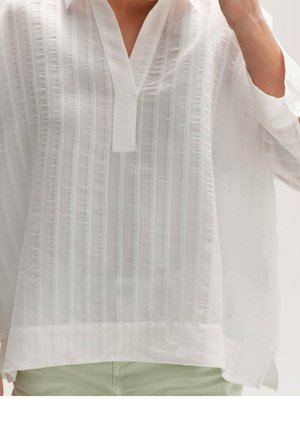 Witte losse blouse met textuur en verticale strepen, V-hals, gedragen met lichtgroene broek door een persoon met opgestoken arm.