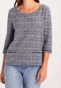 Navy- und weiße strukturierte Strickoberteil mit drei Viertelärmeln, rundem Halsausschnitt und Fransenakzenten an zwei oberen Taschen. Lässiger Schnitt.