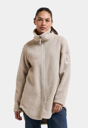 Vrouw draagt een beige sherpa fleece jas met hoge kraag en rits aan de voorkant, staande tegen een effen lichte achtergrond.