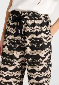 Pantalons à motif noir et beige avec une taille élastique à cordon de serrage et des poches latérales ; le tissu a une texture lisse et un design ondulé.