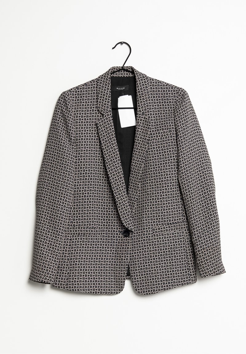 Blazer à carreaux noir et blanc avec un bouton, suspendu sur un cintre noir sur un fond blanc uni.