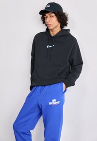 Fekete kapucnis pulóver elülső zsebbel és türkiz Nike logóval, kék melegítőnadrággal, amelyen fehér logós szöveg található. Laid-back sportos viselet.