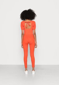 Macacão laranja brilhante com mangas bufantes e um painel traseiro quadrado com um laço. Leggings texturizadas com um padrão subtil, acompanhadas de sapatos brancos.