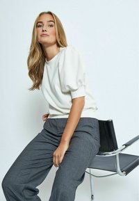 Biely top s balónovými rukávmi vyrobený z mäkkého materiálu, skombinovaný so šedými priliehavými nohavicami, sediaci na elegantnej chrómovo čiernej stoličke.