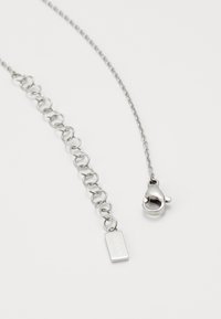 Collier chaîne en argent avec maillons entrelacés, une petite plaque rectangulaire avec un logo embossé, et une fermeture à mousqueton.