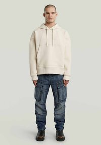 Crème hoodie met een voorzak en trekkoorden, gecombineerd met blauwe cargo jeans met meerdere zakken en een taps toelopende pijp. Zwarte laarzen.