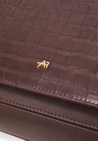 Hnědá kožená taška s krokodýlím vzorem, hladkým povrchem a zlatým kováním s připevněnými písmeny "A7".