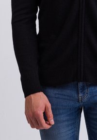 Cardigan nero in maglia con polsini e orlo a coste, caratterizzato da una zip frontale e due tasche laterali, abbinato a jeans in denim blu.