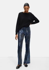 Czarny, oversize sweter w połączeniu z niebieskimi spodniami dzwonowymi ozdobionymi cekinami w pionowe paski; model w czarnych butach, stoi.