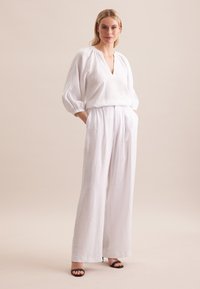 Top in lino bianco con scollatura a V e maniche a palloncino, abbinato a pantaloni larghi in lino bianco. Tacchi neri aperti completano l'outfit.