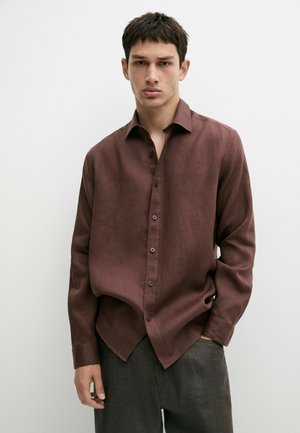 Giovane uomo che indossa una camicia marrone larga con bottoni e pantaloni grigi, in piedi davanti a uno sfondo bianco neutro.