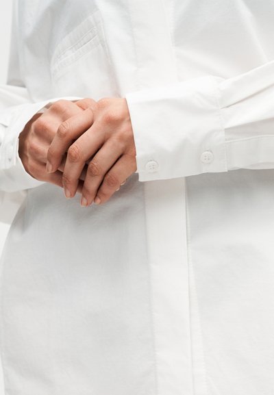 Χέρια σταυρωμένα στη μέση, φορώντας λευκό πουκάμισο με μακριά μανίκια και κουμπιά, με έμφαση στις μανσέτες του πουκάμισου και στον φυσικό τόνο του δέρματος.