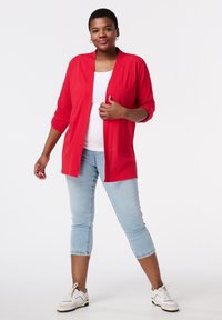 Cardigan rouge à la texture légère, à l'avant ouvert et aux manches trois-quarts, associé à un jean court bleu clair et des baskets blanches.