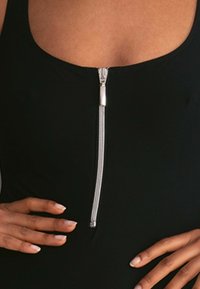Schwarzer Einteiler mit einem Frontreißverschluss, silbernen Beschlägen und einem runden Ausschnitt. Glatte Textur mit taillierter Passform.