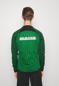Jersey atlético de color verde con un patrón y acentos en negro; presenta motivos de triángulos repetidos y un logo blanco situado en la parte posterior.
