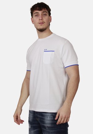 Giovane uomo che indossa una t-shirt bianca a maniche corte con bordo blu sulla tasca e sulle maniche, abbinata a jeans scuri slavati, su sfondo neutro.