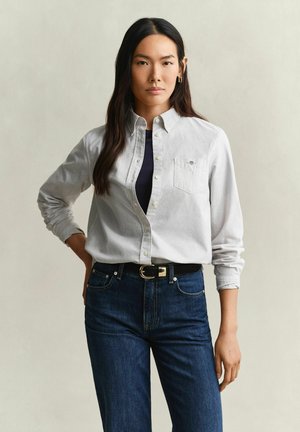 Vrouw met lang donker haar, gekleed in een licht gestreept overhemd met knopen over een zwart topje, blauwe jeans en een zwarte riem, staand met één hand op de heup.