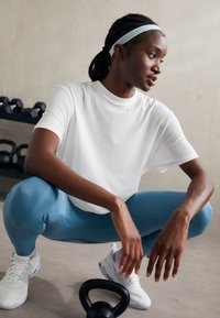 Camiseta branca de manga curta, leggings azuis ajustadas e sapatilhas brancas de desporto. A pessoa está agachada perto de um kettlebell preto.
