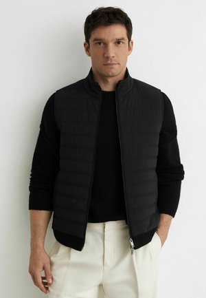 Férfi fekete steppelt mellényben, fekete pulóverrel és törtfehér nadrágban, egy sima fehér fal előtt állva.