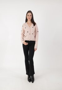 Jasnoróżowy kardigan z czarnymi serduszkami, posiadający dekolt w kształcie litery V i trzy złote guziki, połączony z czarnymi spodniami dzwonami i czarnymi szpilkami.