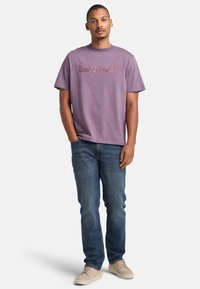 Fialové bavlněné tričko s logem Timberland, spárované s modrými denimovými džíny a béžovými teniskami. Volný střih, krátké rukávy, kulatý výstřih.