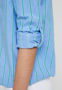 Chemise bouton bleu clair avec des rayures verticales vertes, manches retroussées et un tissu à texture douce. Les accents comprennent un petit bouton aux poignets.