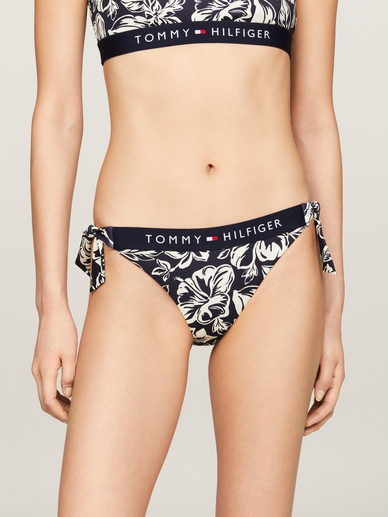 Tommy Hilfiger bikini szett fekete háttérrel, fehér virágmintákkal, állítható oldalsó megkötőkkel és márkázott elasztikus derékkal.