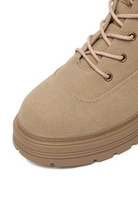 Bocanc de drumeție bej cu un superior din suede texturat, vârf rotund și sireturi de culoare bej deschis. Dispune de găuri metalice pentru șireturi și o talpă din cauciuc stratificată.