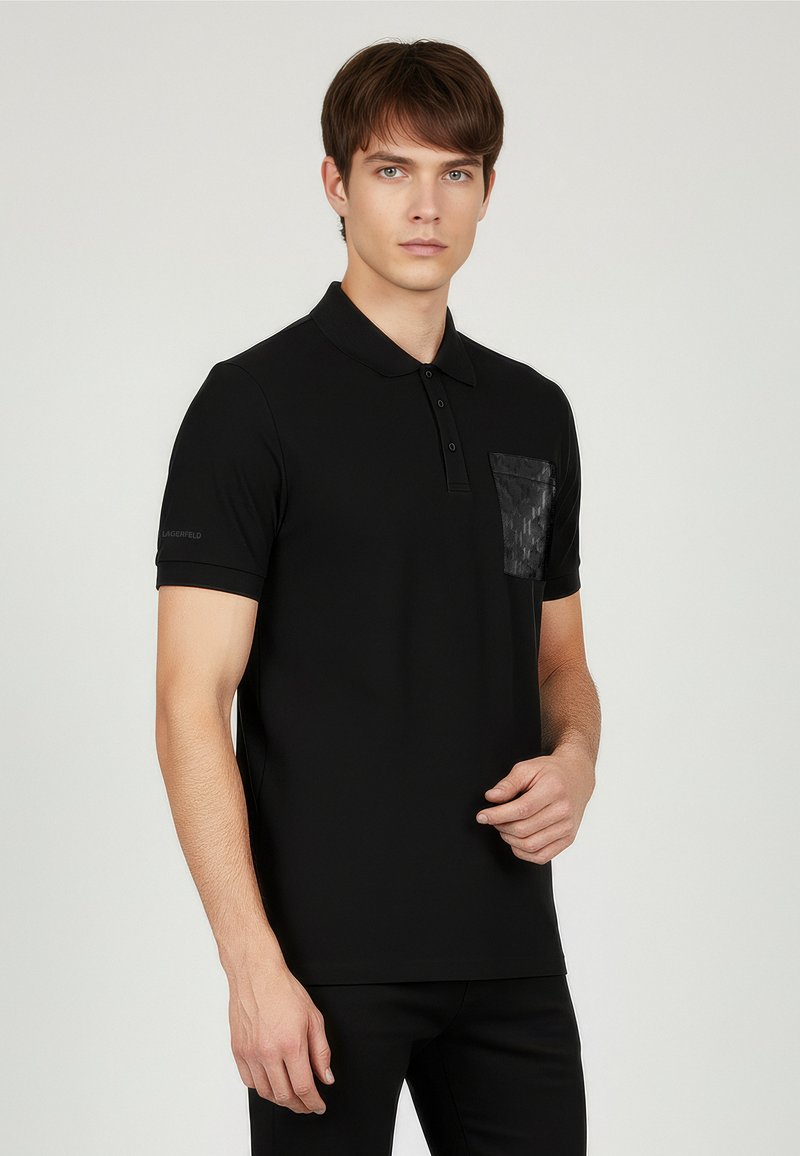 Czarna koszulka polo z teksturowaną, wzorzystą kieszenią. Posiada klasyczny kołnierzyk i krótkie rękawy. Materiał wydaje się gładki i lekki.