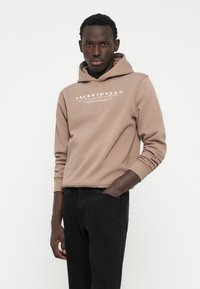 Felpa marrone con cappuccio e lacci, con testo del logo bianco. Abbinata a pantaloni neri, il modello sta in piedi con una mano in tasca.