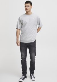 Grå kortermet T-skjorte med "A.R.C." trykk, kombinert med mørke jeans og svarte høye sneakers, stående mot en minimalistisk bakgrunn.