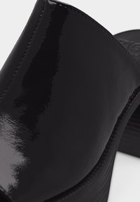 Μαύρο παπούτσι mule από δέρμα πατέντας με χαμηλό τετράγωνο τακούνι και λεία, γυαλιστερή επιφάνεια, σε κοντινή λεπτομέρεια.