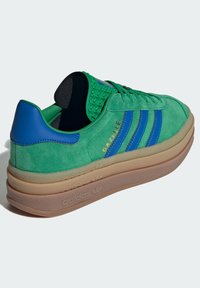 Sapatilhas de camurça verdes com detalhes azuis, apresentando três riscas azuis, uma língua texturizada e uma sola de borracha bege. Branding "Gazelle" em dourado.