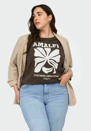 Woman wearing beige blazer over black "Amalfi Costiera Amalfitna Italy" graphic t-shirt and light blue jeans, posing with hand in pocket.