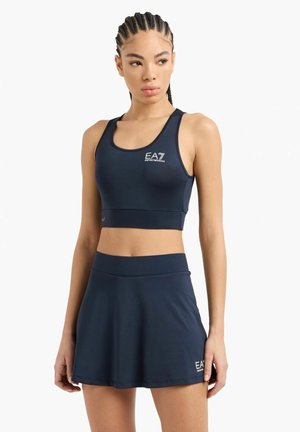 Donna con capelli intrecciati che indossa un top sportivo corto EA7 blu navy e una gonna abbinata, in piedi su uno sfondo bianco.