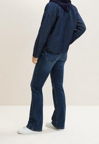 Džínová bunda s dlouhými rukávy, tmavě modré barvy, se skládacími knoflíky, v kombinaci s rozšířenými tmavě modrými džíny a bílými teniskami.