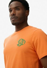Camiseta de algodón naranja con cuello redondo y mangas cortas. Presenta un logo bordado verde en el lado izquierdo del pecho. Textura suave.