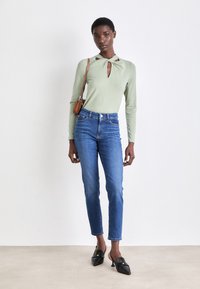 Lys grønne langermede topp med et nøkkelhullskutt, kombinert med tettsittende blå jeans. Svarte slip-on sko med spiss tå og dekorasjon.