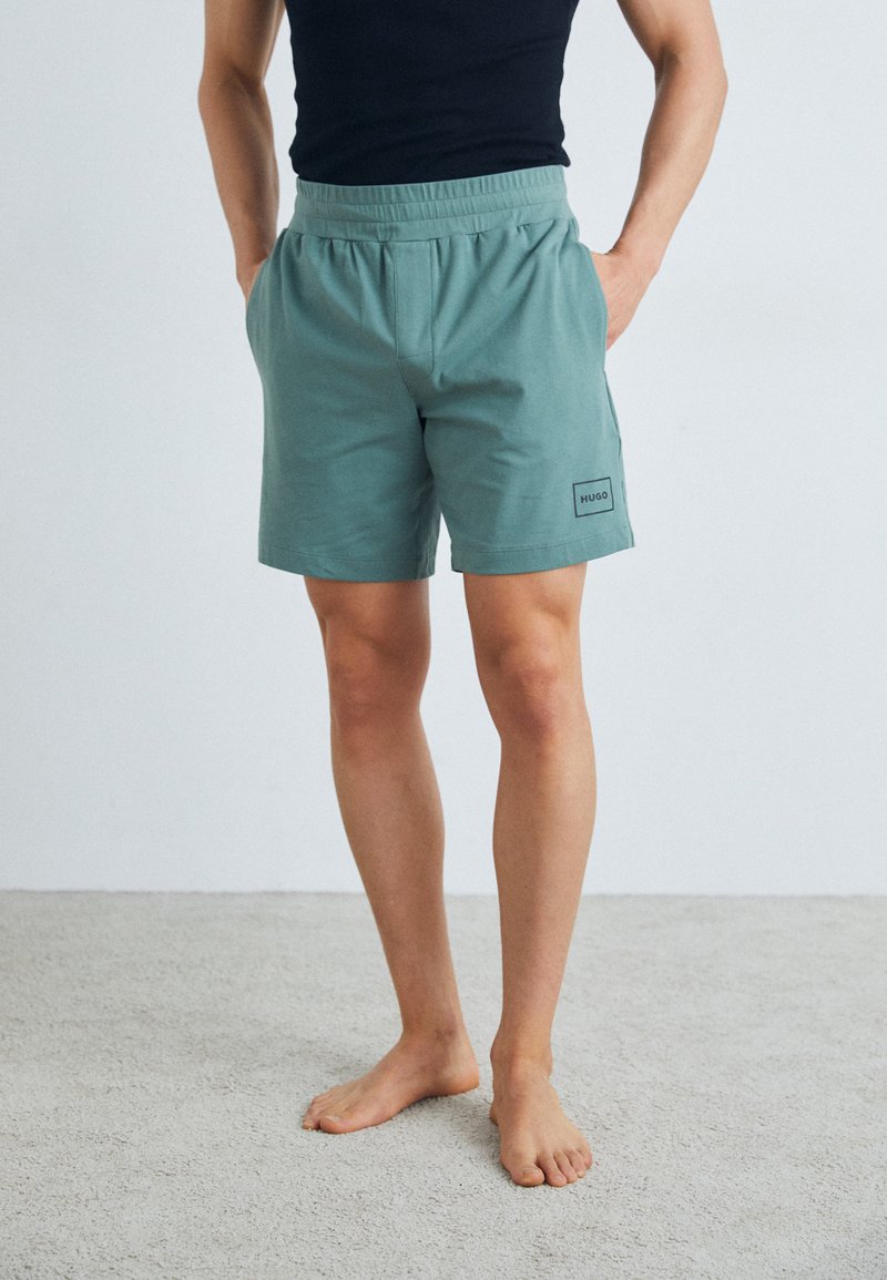 Shorts bleu-vert clair avec une taille élastique et des poches latérales. Présente un petit patch de logo noir sur la cuisse gauche. Matière en coton lisse.