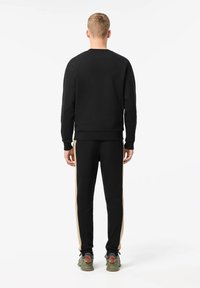 Sweat-shirt noir avec des poignets et un ourlet côtelés, associé à un pantalon noir ajusté avec des rayures dorées sur les côtés. Porté avec des baskets grises et vertes.