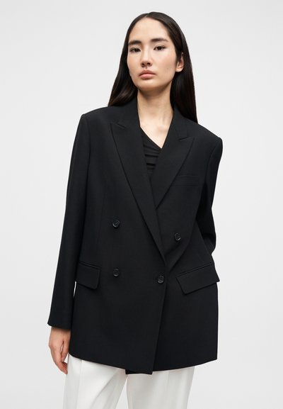 Μαύρο διπλό σακάκι με δομημένους ώμους, δύο εμπρόσθιες τσέπες και διακοσμητικά κουμπιά· κατασκευασμένο από λείο ύφασμα με εφαρμοστή γραμμή.