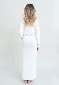 Långärmad vit klänning med en figurnära bodice och flytande kjol, med en åtsittande midja och sidofickor. Ett par beige klackar är synliga.