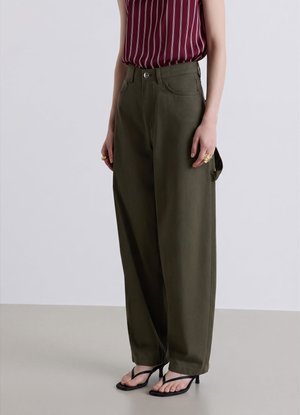 Donna che indossa pantaloni a gamba larga verde oliva a vita alta, top senza maniche a righe bordeaux, sandali neri con tacco e anelli d'oro su entrambe le mani.